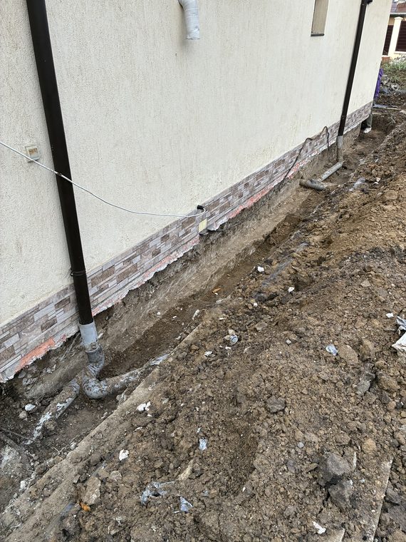 Săpătură de-a lungul fundației unei case locuite, cu placajul soclului desprins și burlanele racordate la țevi noi